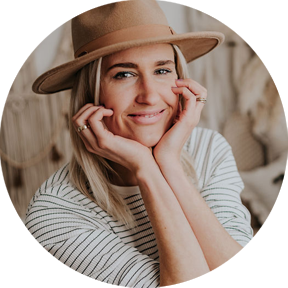 Blonde Frau mit Hut, gestreiftem Oberteil, stützt das Gesicht in die Hände und lächelt